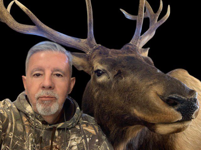 A man posing with a mounted elk head against a dark background.